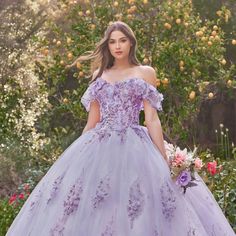 a woman in a purple ball gown standing outside with flowers on the ground and trees behind her