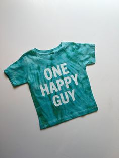 Celebrate your little one's big milestone with a touch of retro charm with our "One Happy Guy" first birthday shirt for baby boys. This tie-dye shirt is the perfect blend of fun and style for your little man's special day. Featuring a vibrant color palette and a playful retro theme, this 1st birthday boy shirt is sure to stand out in all the birthday photos and create lasting memories. Made for comfort and cuteness, this one-year-old boy shirt is a must-have birthday outfit for your little guy a Playful Green T-shirt For Birthday, Green Graphic Print T-shirt For First Birthday, Green T-shirt With Letter Print For First Birthday, Blue Short Sleeve T-shirt For First Birthday, Fun Blue Top For First Birthday, Fun Cotton Tops For First Birthday, Green Cotton T-shirt For First Birthday, Cotton Fun Shirt For First Birthday, Fun Cotton Shirt For First Birthday