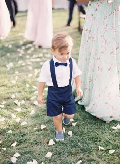 a little boy in a bow tie and suspenders walking towards the camera with other people behind him