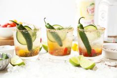 three glasses filled with different types of drinks and garnished with limes, tomatoes, cucumbers, and peppers