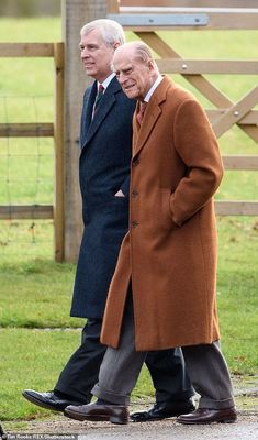 two older men walking down the street in front of a wooden fence and green grass