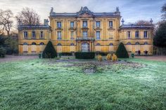 an old building with hedges in front of it on a green grass covered field next to trees