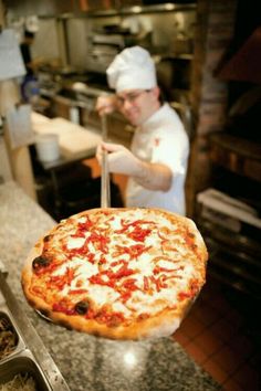 a man holding up a large pizza with toppings on it in a restaurant kitchen