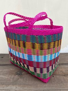 a pink basket with multicolored stripes on the bottom and handles is sitting on a wooden surface