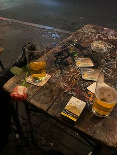 two glasses of beer sit on an old wooden table with books and papers scattered about it