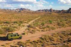 a green vehicle driving down a dirt road in the middle of desert with mountains in the background