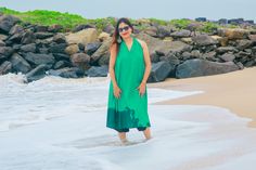 a woman in a green dress standing on the beach next to some rocks and water