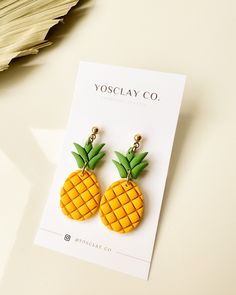 a pair of pineapple shaped earrings on top of a white card next to a plant