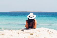 a woman sitting on top of a sandy beach next to the ocean wearing a white hat