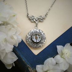 a necklace with a clock on it sitting next to white flowers and a blue book