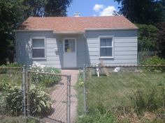 a small gray house behind a chain link fence with a dog in the front yard