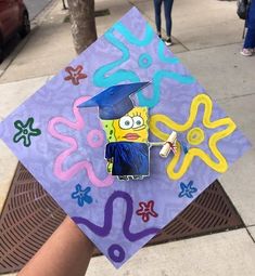 a hand holding a purple and yellow graduation cap with spongebob drawn on it