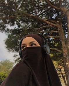 a woman wearing headphones and a brown hijab is standing in front of a tree