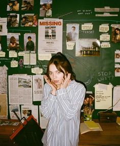 a woman standing in front of a green wall covered with posters and papers, holding her hands to her face