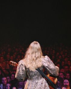 a woman with long blonde hair playing guitar in front of an auditorium full of people