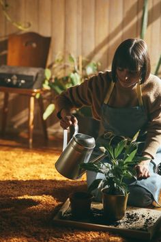 a woman in an apron is watering a potted plant on the floor with a metal watering can