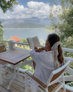 a woman sitting at a table on top of a wooden deck reading a book with mountains in the background