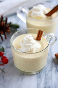 two glasses filled with whipped cream on top of a table next to pine cones and cinnamon sticks