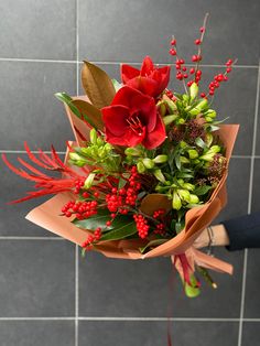 a bouquet of red flowers and greenery is held by someone's hand in front of a tiled wall