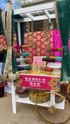 an outdoor vendor selling items for sale on the side of the road with pink and green curtains