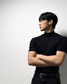 a man standing with his arms crossed wearing a black turtle neck shirt and jeans, against a white wall