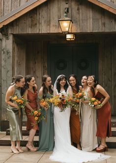 a group of women standing next to each other in front of a wooden building holding bouquets