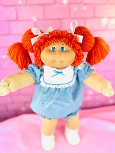 a doll with red hair and blue dress standing in front of a pink brick wall