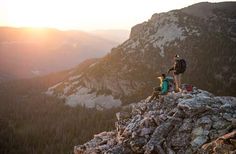 two people standing on top of a mountain with the sun setting behind them and mountains in the background