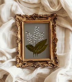 a painting with white flowers in a gold frame on a white cloth covered bed sheet