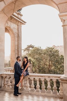 a man and woman standing next to each other on a balcony with an arch in the background