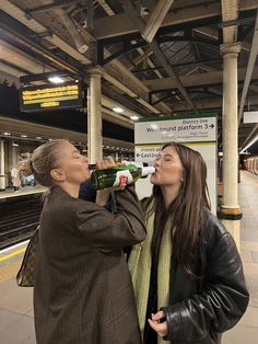 two women standing next to each other at a train station drinking from bottles in their mouths