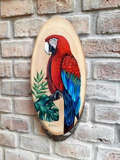 a red and blue parrot painted on a wooden plaque next to a brick wall with green leaves