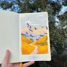 a hand holding up an open book with a painting on the cover and mountains in the background