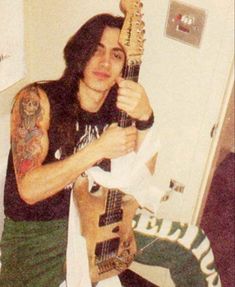 a man with long hair and tattoos holding a guitar in his right hand while posing for the camera