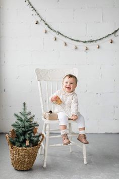 a baby sitting in a white chair next to a small christmas tree and a potted plant