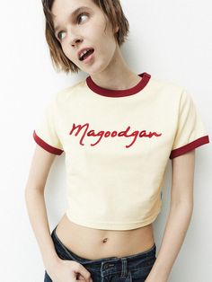 The clothing item displayed is a cropped T-shirt with a casual, sporty design. It features contrasting trim at the collar and sleeve edges, giving it a retro athletic appearance. The fabric appears soft and lightweight, suitable for warm weather or layering. A distinctive cursive script is prominently placed in the center of the chest area, adding a personalized touch to the garment.- The shirt has a ribbed neckline and sleeve hems, creating a finished look and adding to its durability.- The cut Retro Cotton Tops With Contrast Color, Retro Tops With Contrast Color For Spring, Retro Contrast Color Tops For Spring, Retro Cotton Tops With Embroidered Logo, Trendy Crew Neck Top With Contrast Stitching, Cotton Tops With Letter Embroidery In College Style, Cotton College Style Top With Letter Embroidery, Retro Cotton Top With Embroidered Logo, Cotton Retro Crop Top For Streetwear