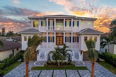 a large white house with blue shutters and palm trees in the front yard at sunset