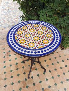 a mosaic table sitting on top of a tiled floor next to a tree and bushes