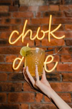 a person holding a drink in front of a brick wall with neon signs above it