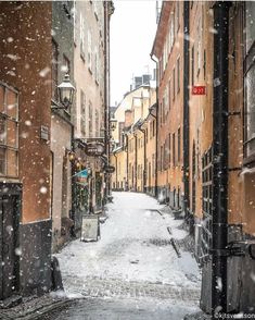 an alley way with snow falling on the ground and buildings lining both sides, in winter