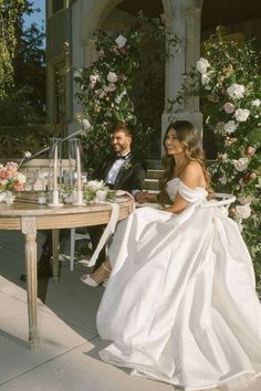 a bride and groom sitting at a table in front of a flower covered building with greenery