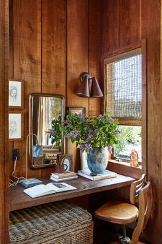 a desk with a mirror, lamp and vase on it next to a window in a wood paneled room