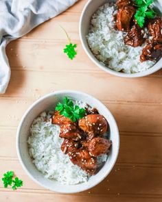 two white bowls filled with rice and meat on top of a wooden table next to green leafy garnishes