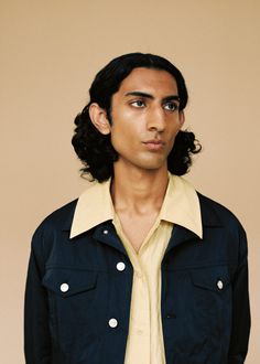 a man with long black hair wearing a blue jacket and yellow shirt is looking at the camera