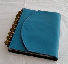 a blue leather notebook with gold spirals on the front and inside cover is laying on a white sheet