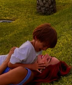 a woman laying on top of a grass covered field next to a little boy with red hair