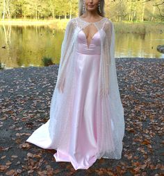 a woman in a pink gown and white cape standing on the ground next to a lake