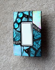 a decorative light switch cover with blue and green designs on the front, sitting on a carpeted floor