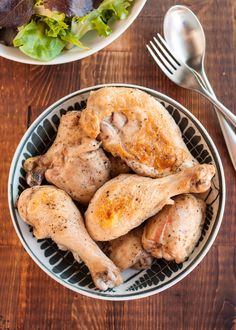 a bowl full of chicken next to a salad on a wooden table with utensils