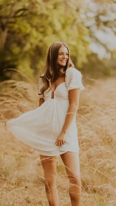 a woman in a white dress is standing in tall grass and smiling at the camera